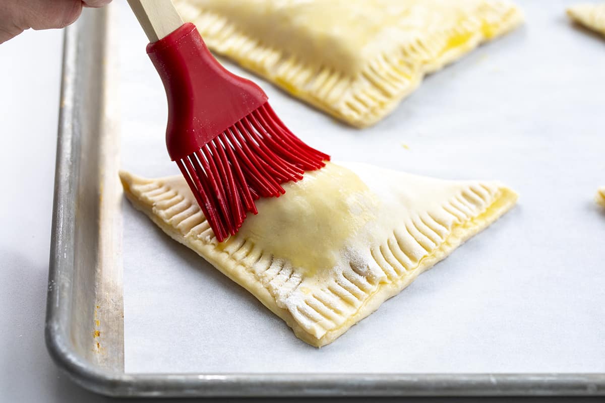 Easy & Amazing Apple Turnovers are filled with spiced apples and baked in flaky, buttery puff pastry dough! Brushing Egg Wash over unbaked Apple Turnovers.