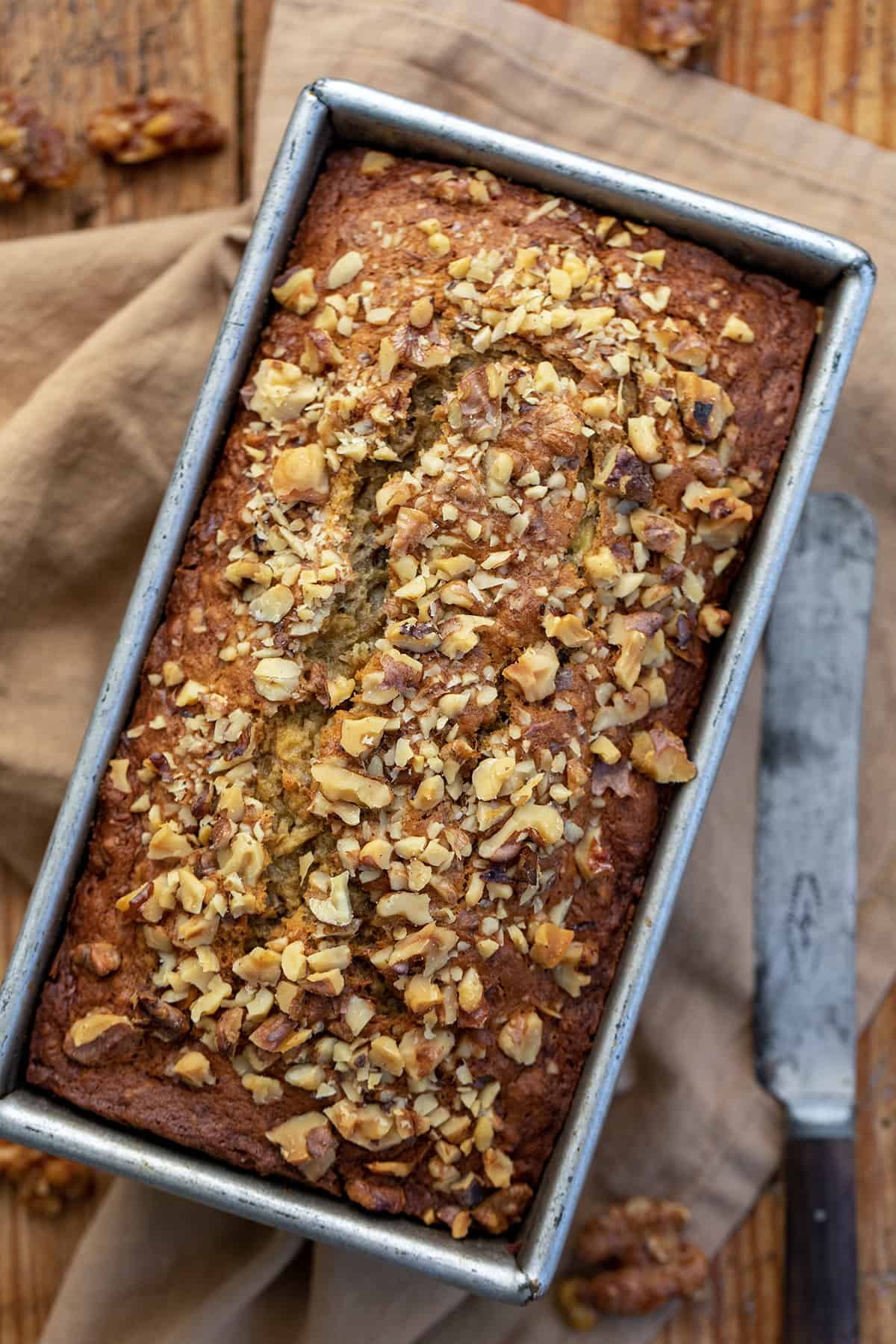 Browned Butter Banana Nut Bread is made by browning butter to add a toasty taste and then loading and topping the bread with chopped walnuts to add even more nutty flavor. It is definitely a step up from traditional banana bread and possibly my new favorite way to enjoy it! (If you prefer a recipe without nuts, I also have my Best Banana Bread Recipe you will want to try.) Looking Down Onto Brown Butter Banana Nut Bread on a Cutting Board with a Knife.