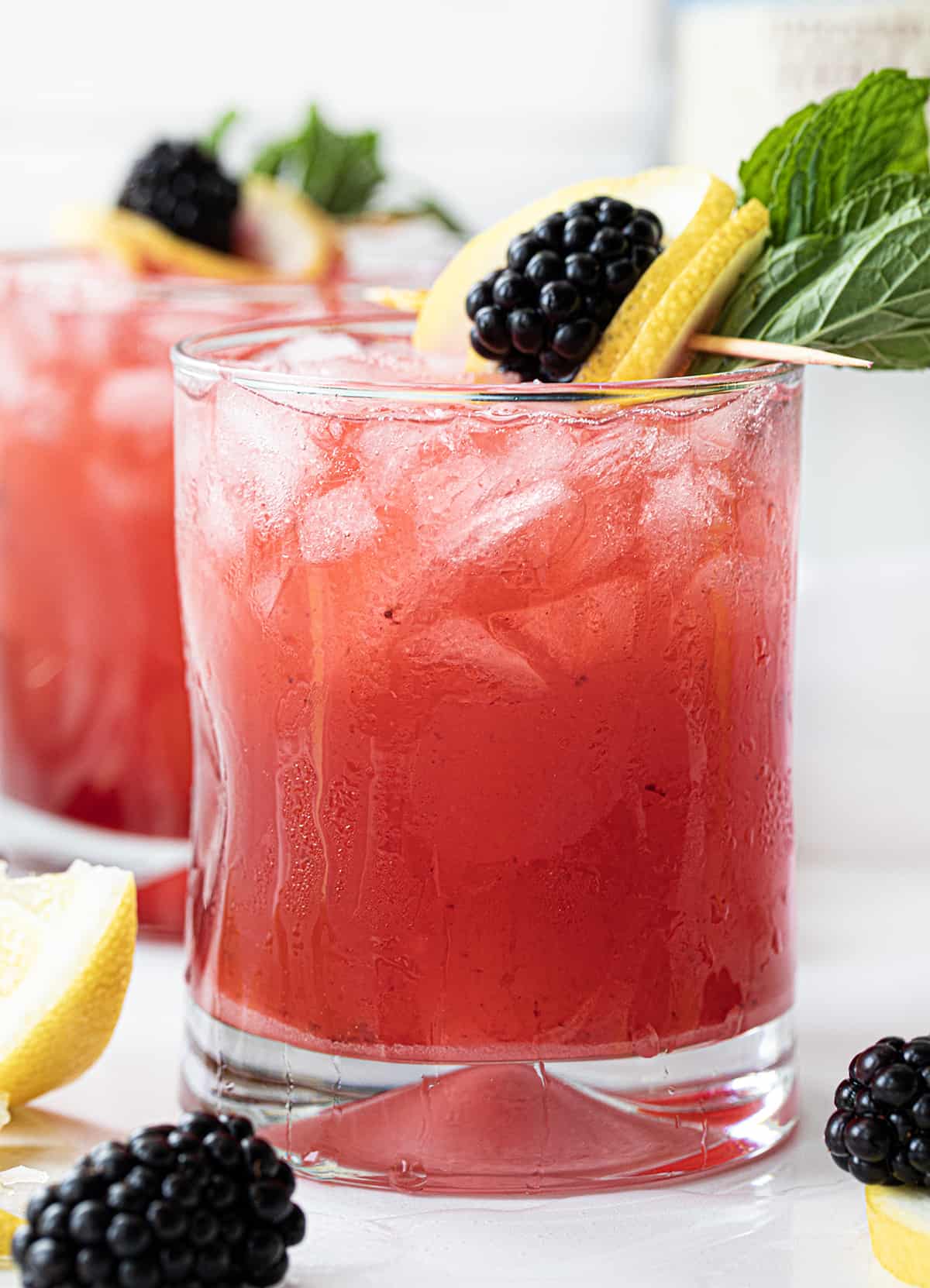 Blackberry Fizz is a refreshing cocktail made with blackberries, freshly squeezed lemon juice, vodka, simple syrup, and club soda. It's just like a blackberry bramble but without blackberry liqueur. It offers a delightful combination of fruity, tangy, and refreshing flavors, making it a perfect choice for a summer sip or any time you're in the mood for a vibrant and lively beverage! Blackberry Fizz Cocktails on a White Counter with Blackberry Lemon Garnish.
