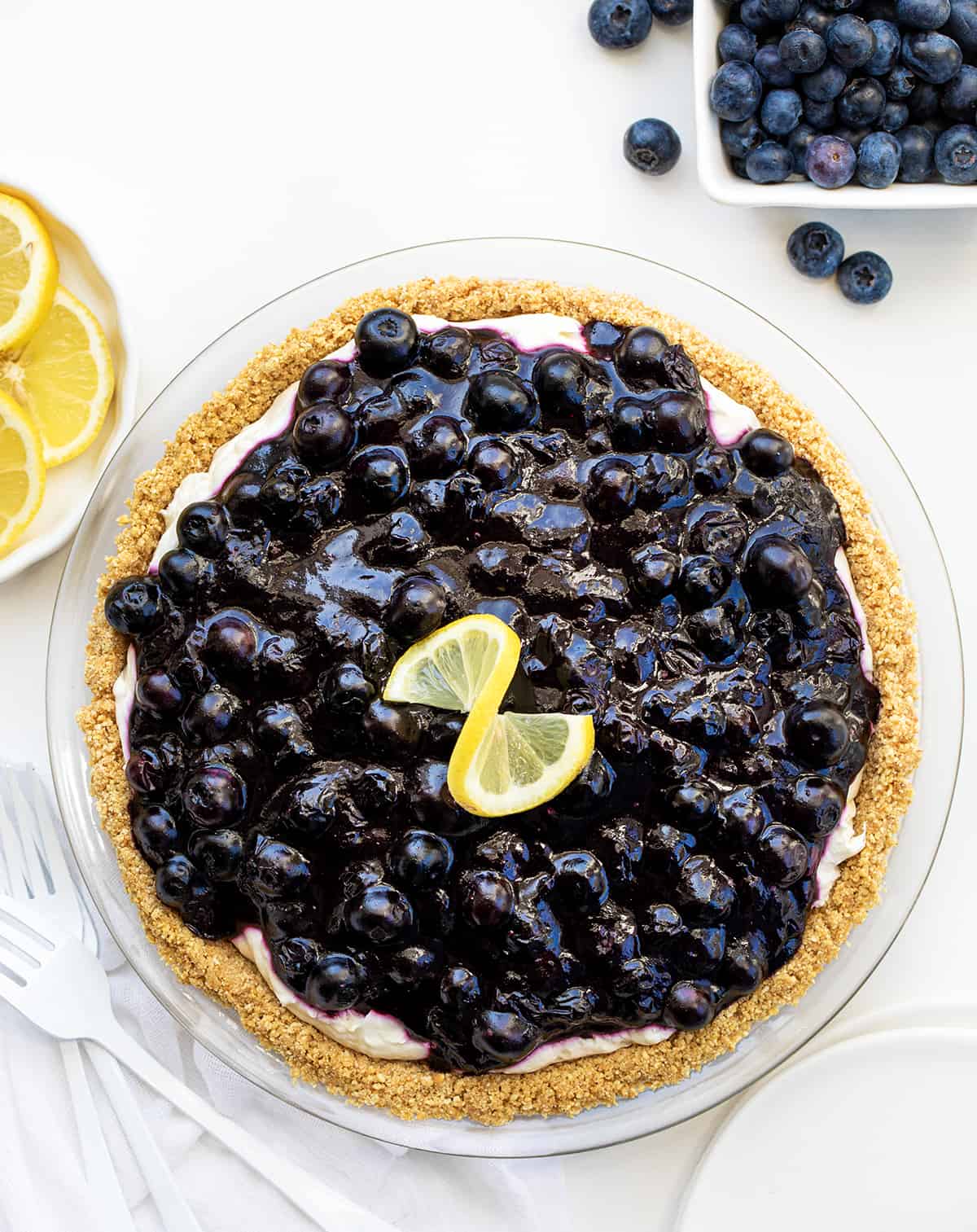 Blueberry Lemon Cream Pie is an ultra-creamy no-bake dessert made with a lemon cheesecake filling and homemade blueberry topping, all in a graham cracker crust. The tangy, silky-smooth lemon filling complements the sweetness of the blueberry sauce creating a luscious treat! Looking Down on a Blueberry Lemon Cream Pie on a White Counter Surrounded by Blueberries and Lemons and White Forks.