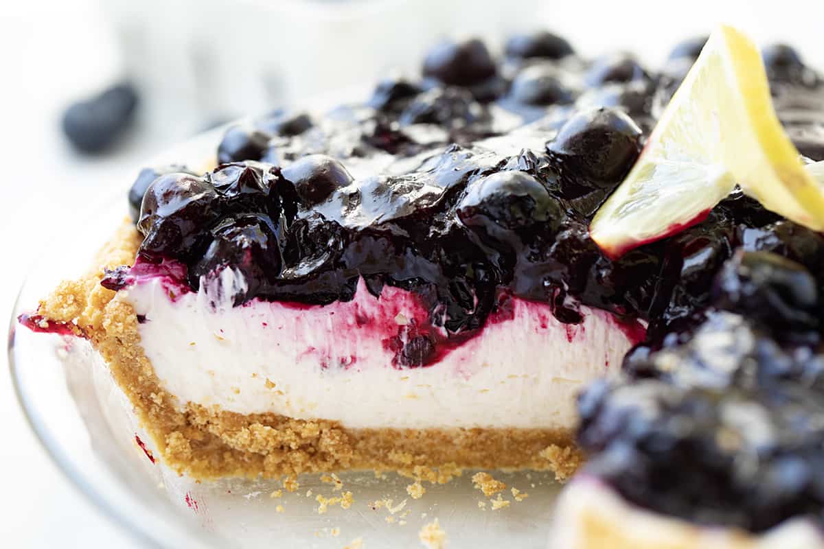 Blueberry Lemon Cream Pie is an ultra-creamy no-bake dessert made with a lemon cheesecake filling and homemade blueberry topping, all in a graham cracker crust. The tangy, silky-smooth lemon filling complements the sweetness of the blueberry sauce creating a luscious treat! Inside of Blueberry Lemon Cream Pie Still in Pie Plate with a Piece Removed Showing Inside.