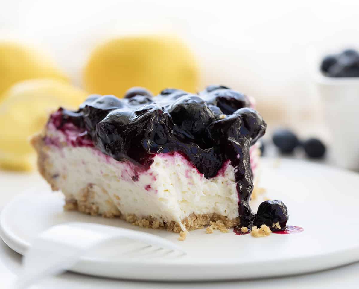Blueberry Lemon Cream Pie is an ultra-creamy no-bake dessert made with a lemon cheesecake filling and homemade blueberry topping, all in a graham cracker crust. The tangy, silky-smooth lemon filling complements the sweetness of the blueberry sauce creating a luscious treat! Piece of Blueberry Lemon Cream Pie on a White Plate With a Bite Removed.