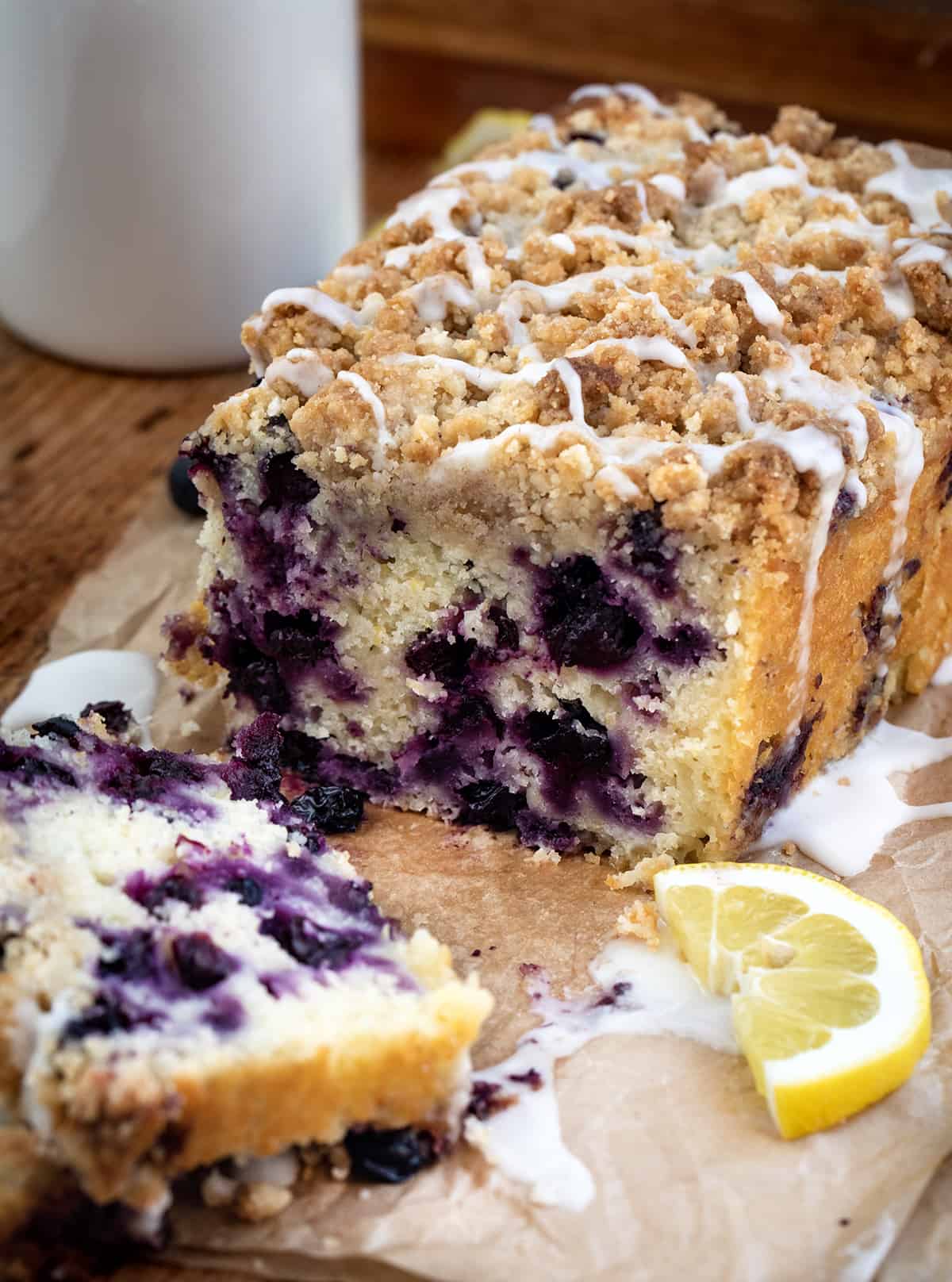 Blueberry Lemon Loaf is a moist cake-like loaf loaded with fresh blueberries and lemon flavor, topped and baked with a crumble topping. Then, the loaf is drizzled with a sweet glaze to make this a perfect breakfast, snack, or dessert. This is a copycat of the viral Costco Blueberry Lemon Loaf and dare I say, even BETTER!! Cut Into Blueberry Lemon Loaf with Glaze and Lemons on Parchment on a Cutting Board.