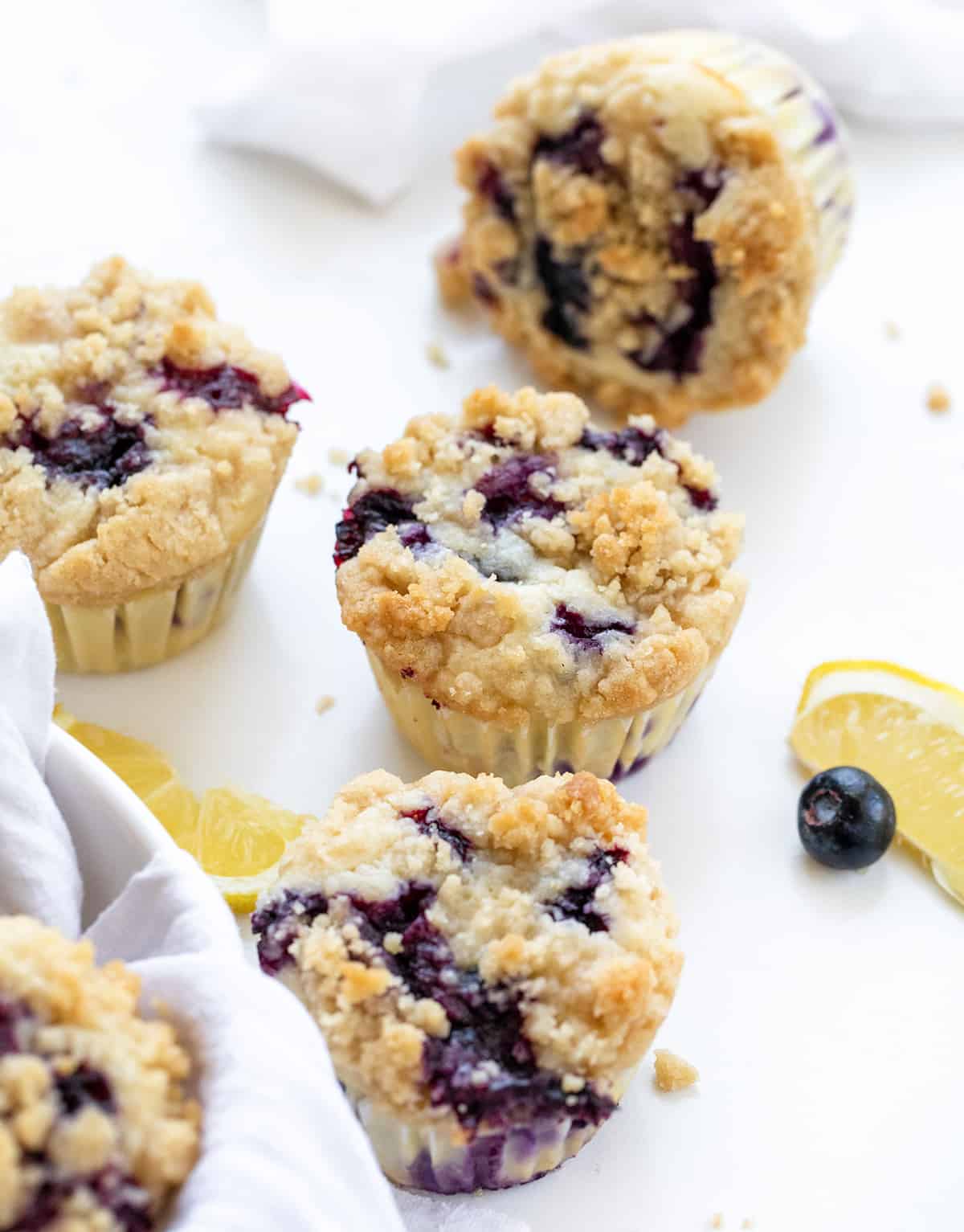Blueberry Lemon Crumb Muffins are moist and tender muffins loaded with fresh blueberries and lemon flavor, topped and baked with a crispy, buttery crumble. They are the perfect sweet snack, dessert, or breakfast! Blueberry Lemon Muffins on a White Counter with Fresh Lemon Slice and Blueberries.