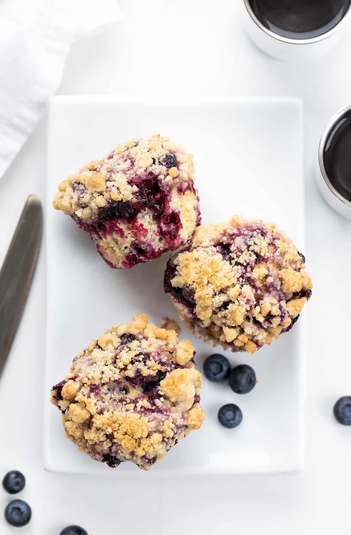 Blueberry Muffins are sweet and fluffy muffins made with fresh blueberries, homemade blueberry syrup, and a crumble topping. Double the blueberry! They are a delicious breakfast, snack, or treat to enjoy with a cup of coffee or tea and a great way to use up ripe blueberries! Blueberry Muffins on a White Plate with Blueberries around them.