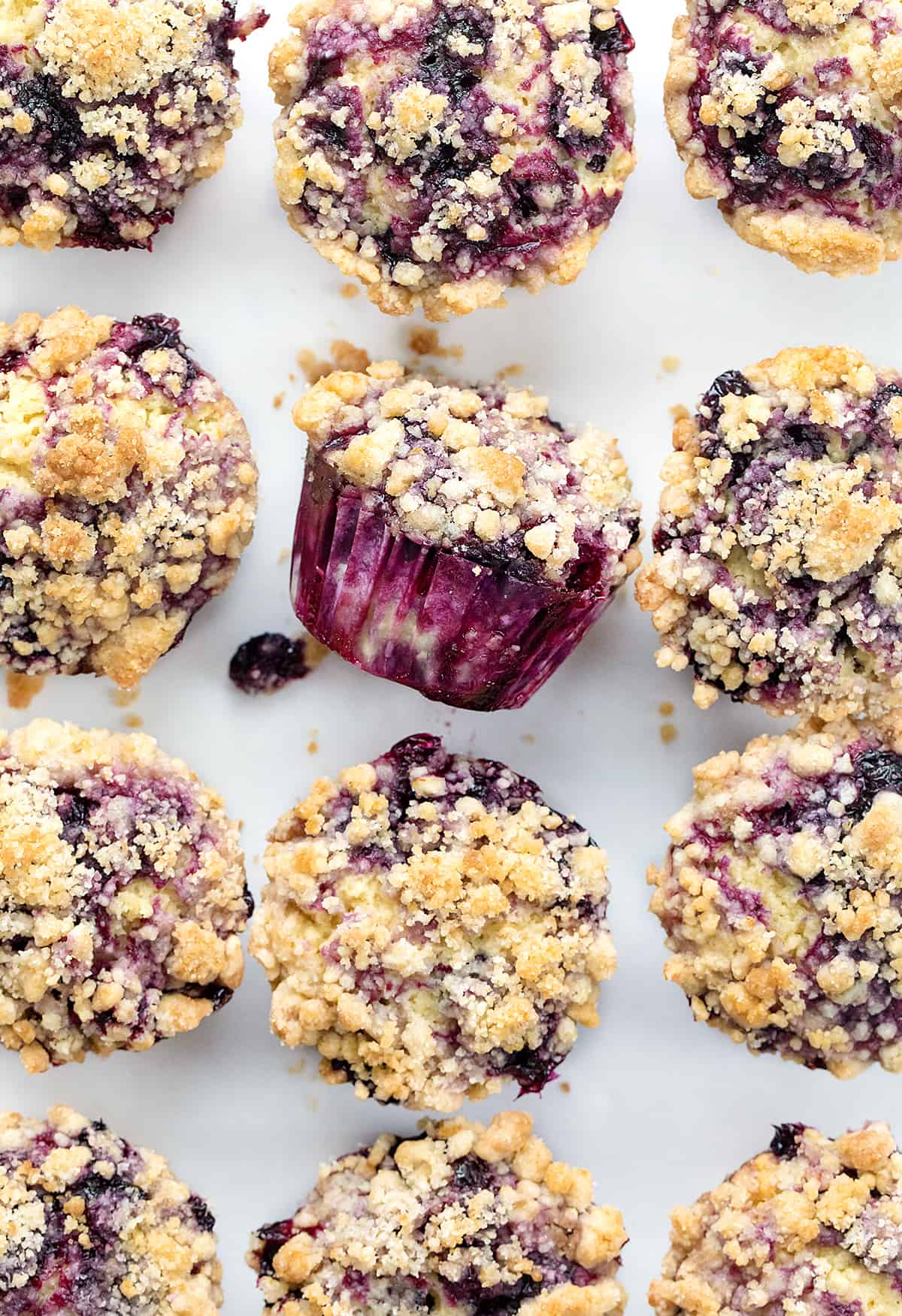 Blueberry Muffins are sweet and fluffy muffins made with fresh blueberries, homemade blueberry syrup, and a crumble topping. Double the blueberry! They are a delicious breakfast, snack, or treat to enjoy with a cup of coffee or tea and a great way to use up ripe blueberries! Blueberry Muffins on a Counter with One Tipped on Its Side.