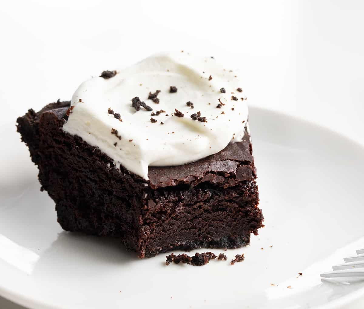 Brownie Pie combines the irresistible textures of a crisp Oreo cookie crust, dense and fudgy brownies, and fluffy whipped cream with a chocolate drizzle. It's a perfect dessert for special occasions or anytime you're craving a truly indulgent treat! Piece of Brownie Pie on a White Plate with Bite Removed Showing Inside Texture.