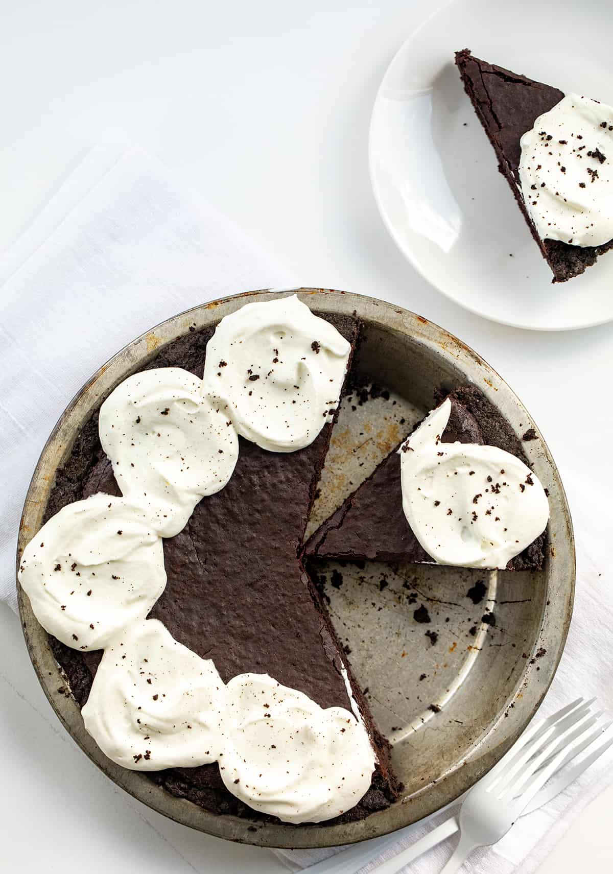 Brownie Pie combines the irresistible textures of a crisp Oreo cookie crust, dense and fudgy brownies, and fluffy whipped cream with a chocolate drizzle. It's a perfect dessert for special occasions or anytime you're craving a truly indulgent treat! Pie Pan with Brownie Pie in it and a Few Pieces Removed on a White Counter.