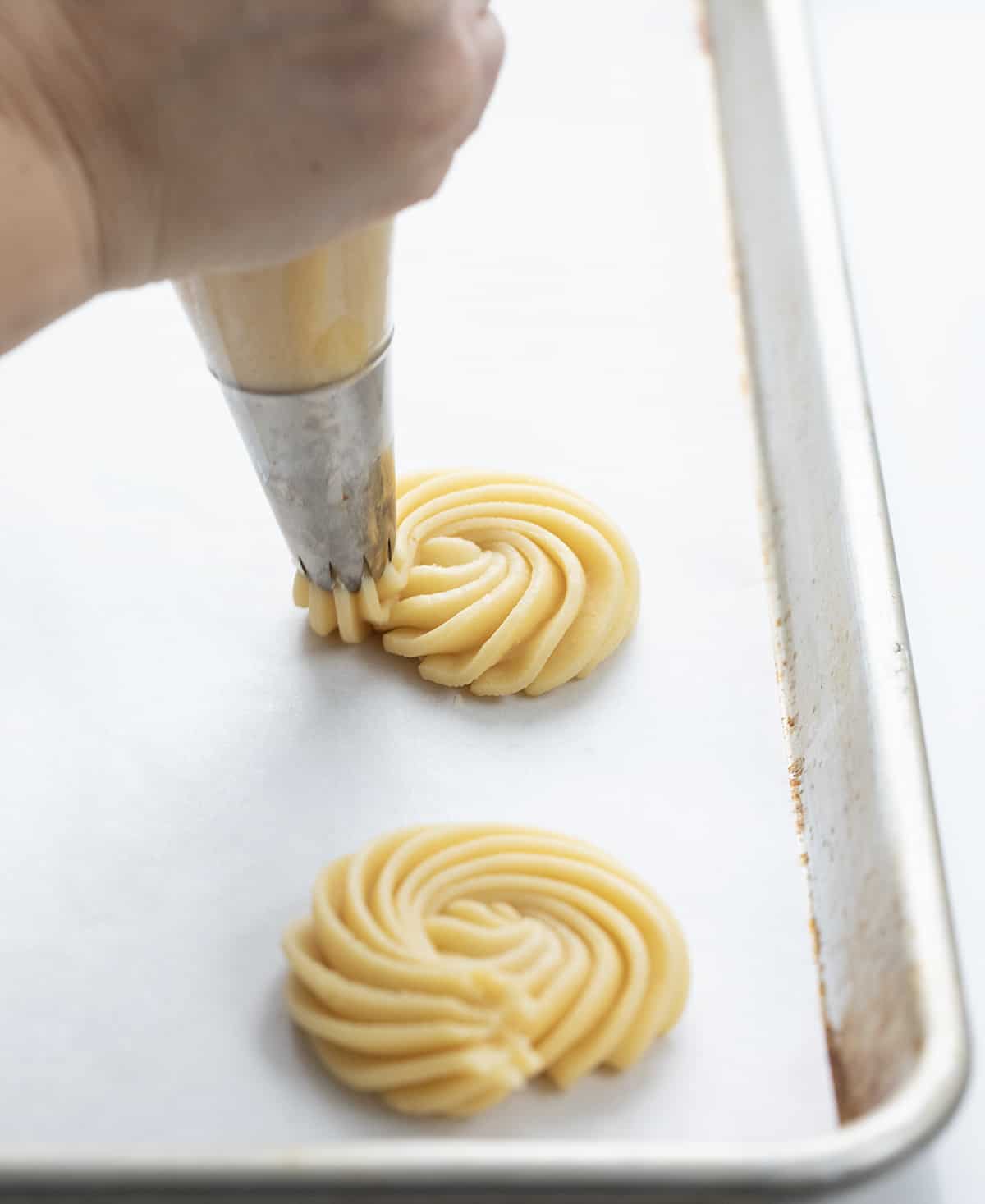Butter Cookies are buttery, tender cookies that can be enjoyed as-is with a cup of coffee or tea or as a base for decorating holiday cookies. They are synonymous with Christmas cookies (remember the blue tin?), and I wanted to find a recipe as easy as possible. I know how busy the holiday season can be, so a cookie recipe that does not take all day with no chilling time works for me (and it will for you)! Perfect for a cookie exchange or a Christmas dessert charcuterie board! Piping a butter cookie.
