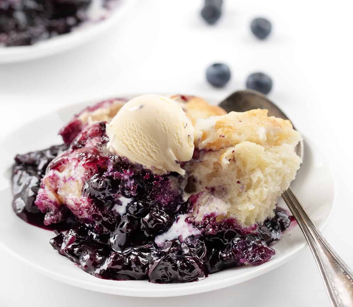 Butter Swim Biscuit Blueberry Cobbler is a delightful dessert that combines homemade blueberry filling with tender, buttery biscuits. It's a perfect treat for gatherings, family meals, or anytime you crave a comforting and fruity dessert. It is so easy to throw together and ready in 30 minutes! Plate of Butter Swim Biscuit Blueberry Cobbler on a White Counter with Blueberries.