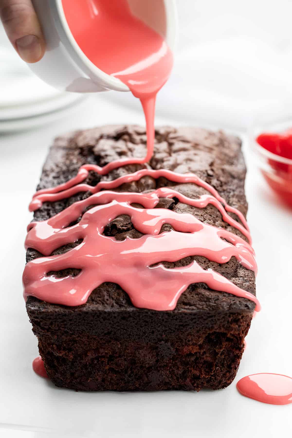 Cherry Brownie Bread is a dessert loaf of bread that merges the irresistible taste of a rich, chocolatey brownie with added cherries, baked, and topped with a cherry glaze. The cherries add a pop of bright flavor and a contrasting texture to the dense, chewy brownie bread. Whether you enjoy it as a sweet snack or as an after-dinner dessert, cherry brownie bread is sure to satisfy your cravings for something sweet and indulgent! Drizzling Cherry Glaze Over Cherry Brownie Bread on a White Counter.