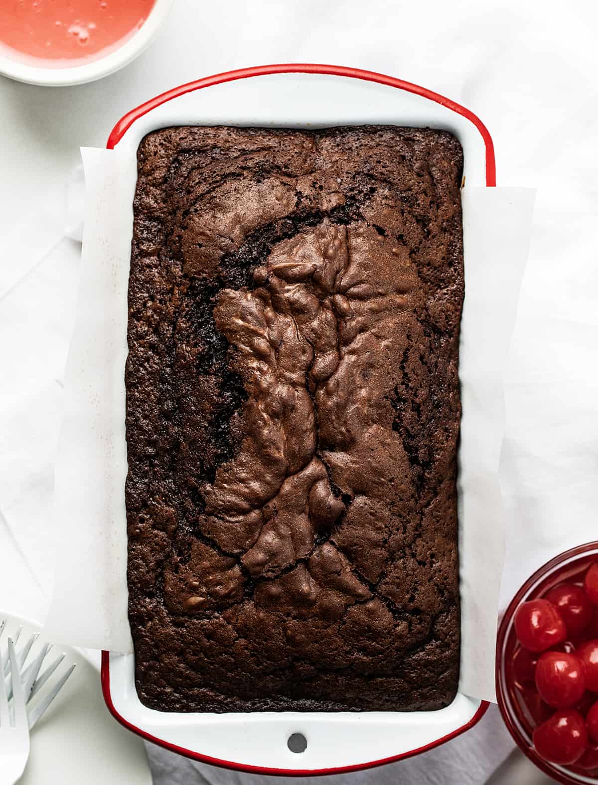 Cherry Brownie Bread is a dessert loaf of bread that merges the irresistible taste of a rich, chocolatey brownie with added cherries, baked, and topped with a cherry glaze. The cherries add a pop of bright flavor and a contrasting texture to the dense, chewy brownie bread. Whether you enjoy it as a sweet snack or as an after-dinner dessert, cherry brownie bread is sure to satisfy your cravings for something sweet and indulgent! Loaf of Cherry Brownie Bread in a White Pan with Red Rim on a White Counter.
