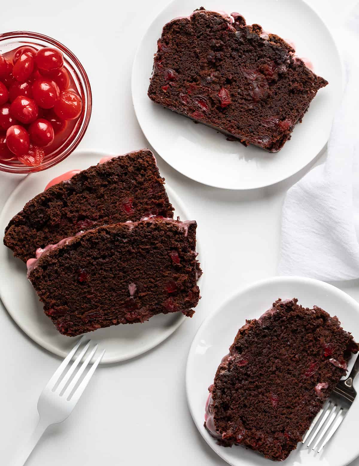 Cherry Brownie Bread is a dessert loaf of bread that merges the irresistible taste of a rich, chocolatey brownie with added cherries, baked, and topped with a cherry glaze. The cherries add a pop of bright flavor and a contrasting texture to the dense, chewy brownie bread. Whether you enjoy it as a sweet snack or as an after-dinner dessert, cherry brownie bread is sure to satisfy your cravings for something sweet and indulgent! Slices of Cherry Brownie Bread on Plates on a White Counter with a Small Bowl of Cherries.
