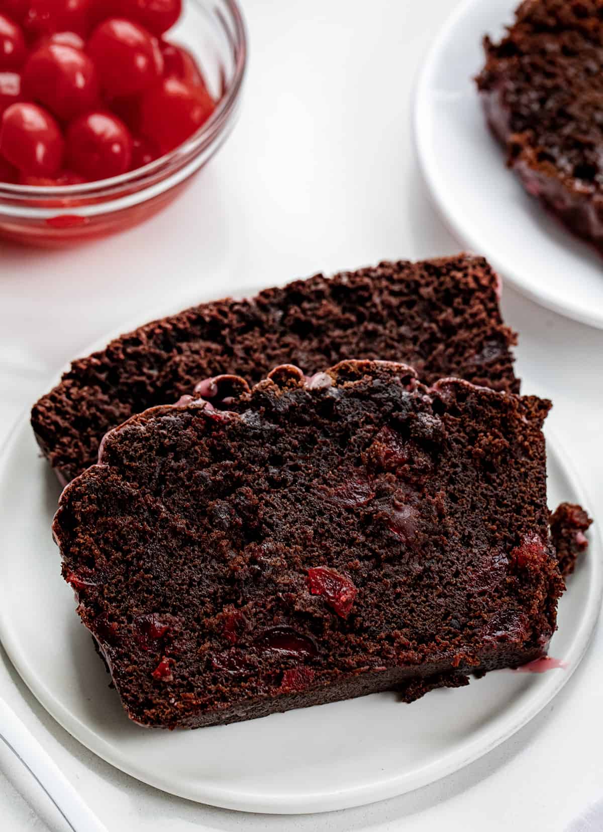 Cherry Brownie Bread is a dessert loaf of bread that merges the irresistible taste of a rich, chocolatey brownie with added cherries, baked, and topped with a cherry glaze. The cherries add a pop of bright flavor and a contrasting texture to the dense, chewy brownie bread. Whether you enjoy it as a sweet snack or as an after-dinner dessert, cherry brownie bread is sure to satisfy your cravings for something sweet and indulgent! Close up of Cherry Brownie Bread Slices on a Plate with Cherries in the Background.