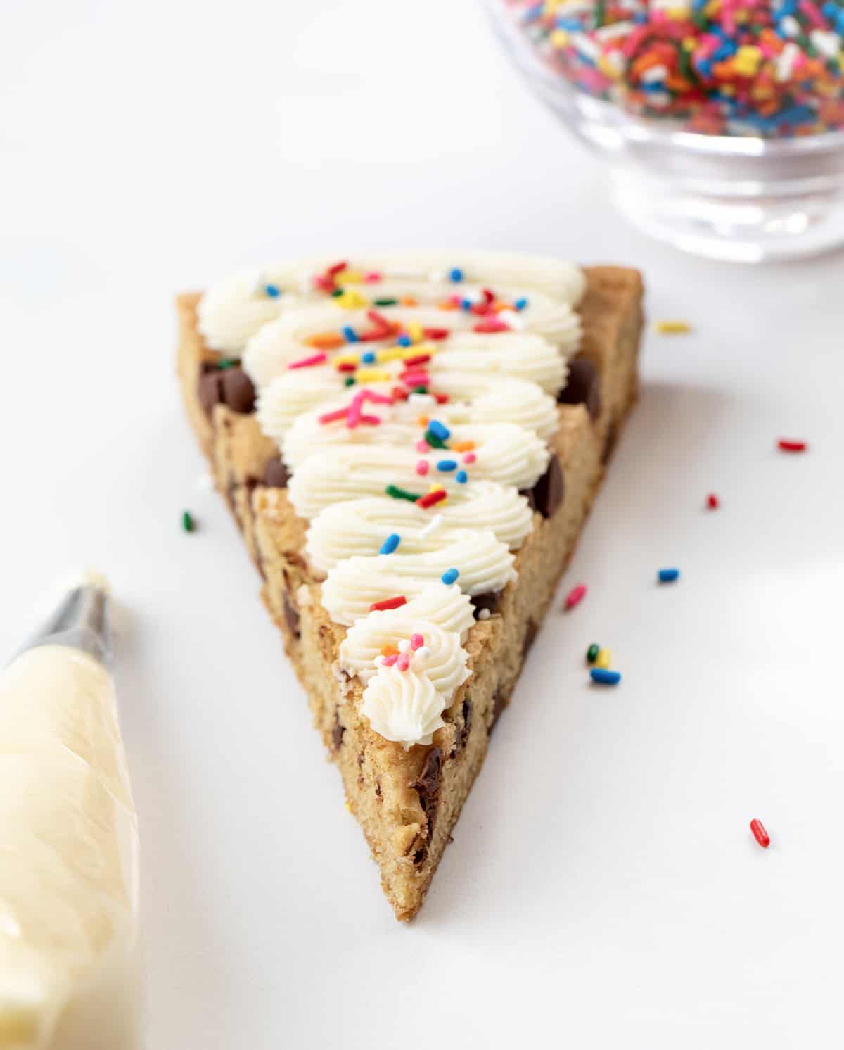 Cookie Cake For A Crowd is a colossal chocolate chip cookie frosted in three sections with cookie dough frosting, whipped chocolate buttercream, and American buttercream. The cookie itself has a soft and tender texture that is delightful on its own. Then, the frosting adds an extra layer of indulgence! It's like three treats in one! One Piece of Cookie Cake for a Crowd with Vanilla Frosting, Sprinkles, and Piping Bag.