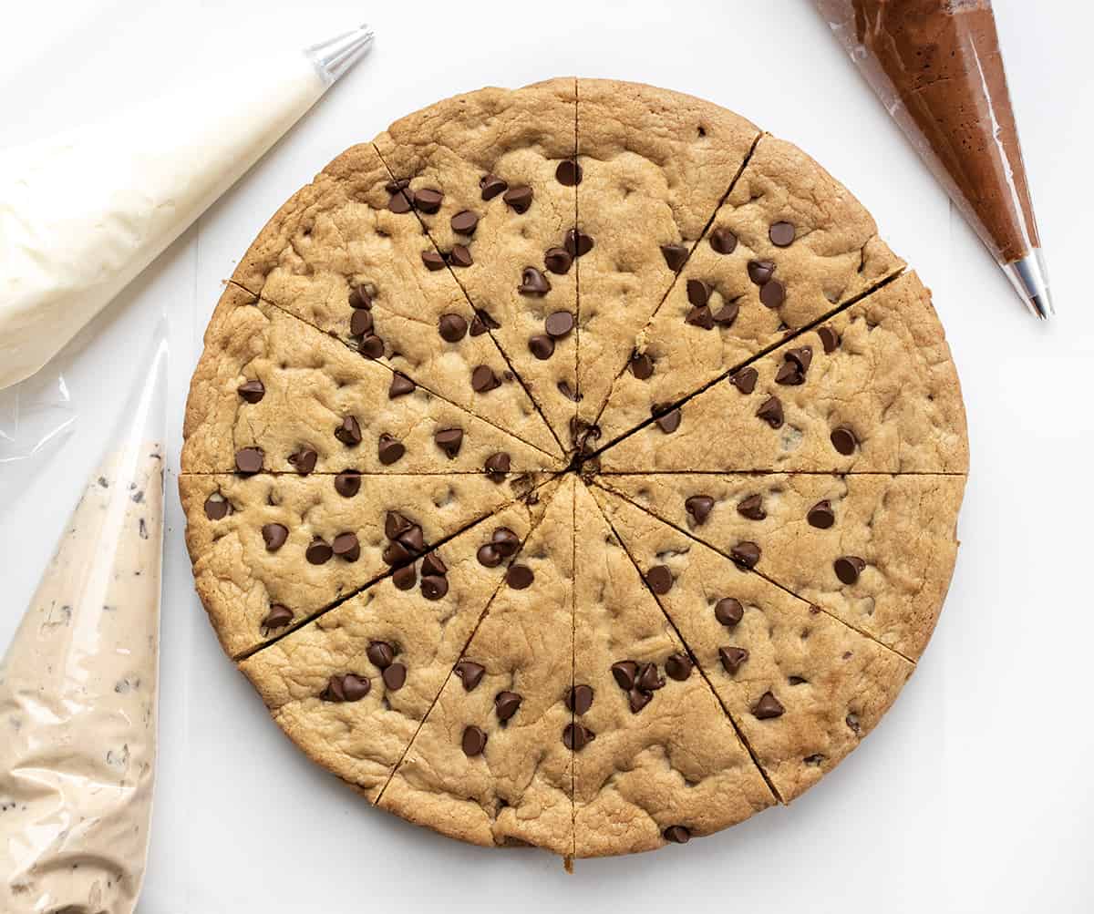 Cookie Cake For A Crowd is a colossal chocolate chip cookie frosted in three sections with cookie dough frosting, whipped chocolate buttercream, and American buttercream. The cookie itself has a soft and tender texture that is delightful on its own. Then, the frosting adds an extra layer of indulgence! It's like three treats in one! Baked Cookie Cake Cut into 12 Pieces with Vanilla, Chocolate, and COokie Dough Frosting in Piping Bags.