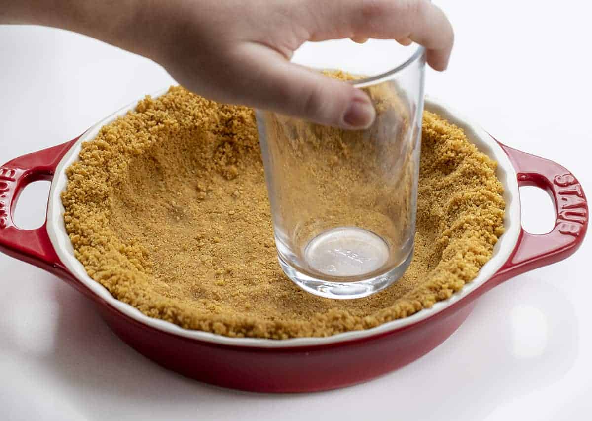 No-Bake Eggnog Pie is a creamy pie made with vanilla pudding mix, nutmeg, eggnog, rum, and whipped cream that tastes just like a glass of eggnog! Pie, No Bake Pie, Eggnog Pie, Leftover Eggnog Recipes, How to Use Eggnog, Graham Cracker Crust Pie, Dessert, Easy Pie, New Years Pie, i am baker, iambaker Hand Pressing Down Graham Cracker Crust with a Glass