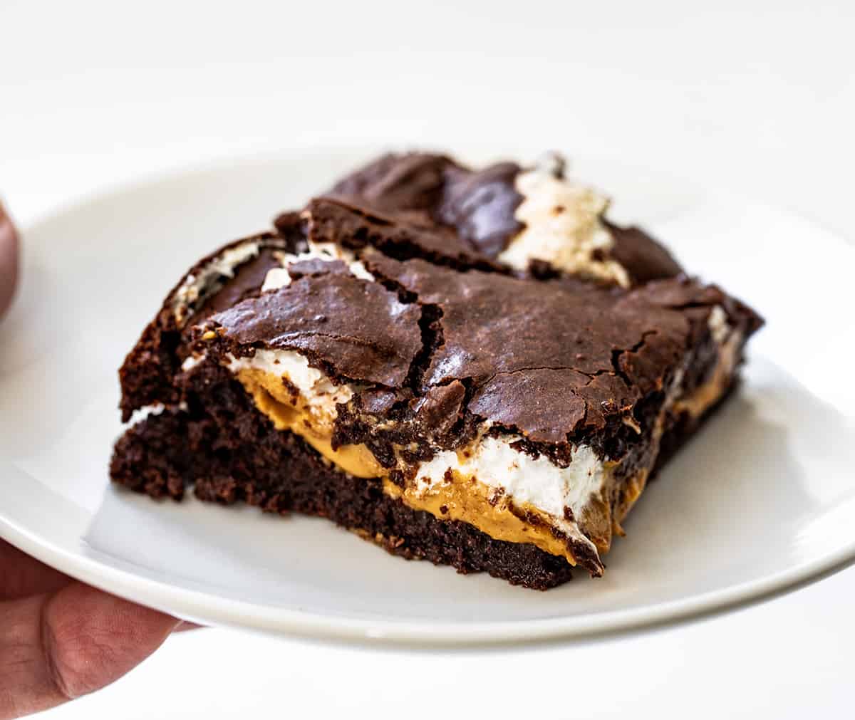 Fluffernutter Brownies are creamy peanut butter and marshmallow fluff sandwiched and baked between two layers of the ultimate fudgy brownies. The brownie layers provide a rich, chocolaty flavor, while the peanut butter and marshmallow layers add a creamy and fluffy contrast! Fluffernutter Brownie on a White Plate.