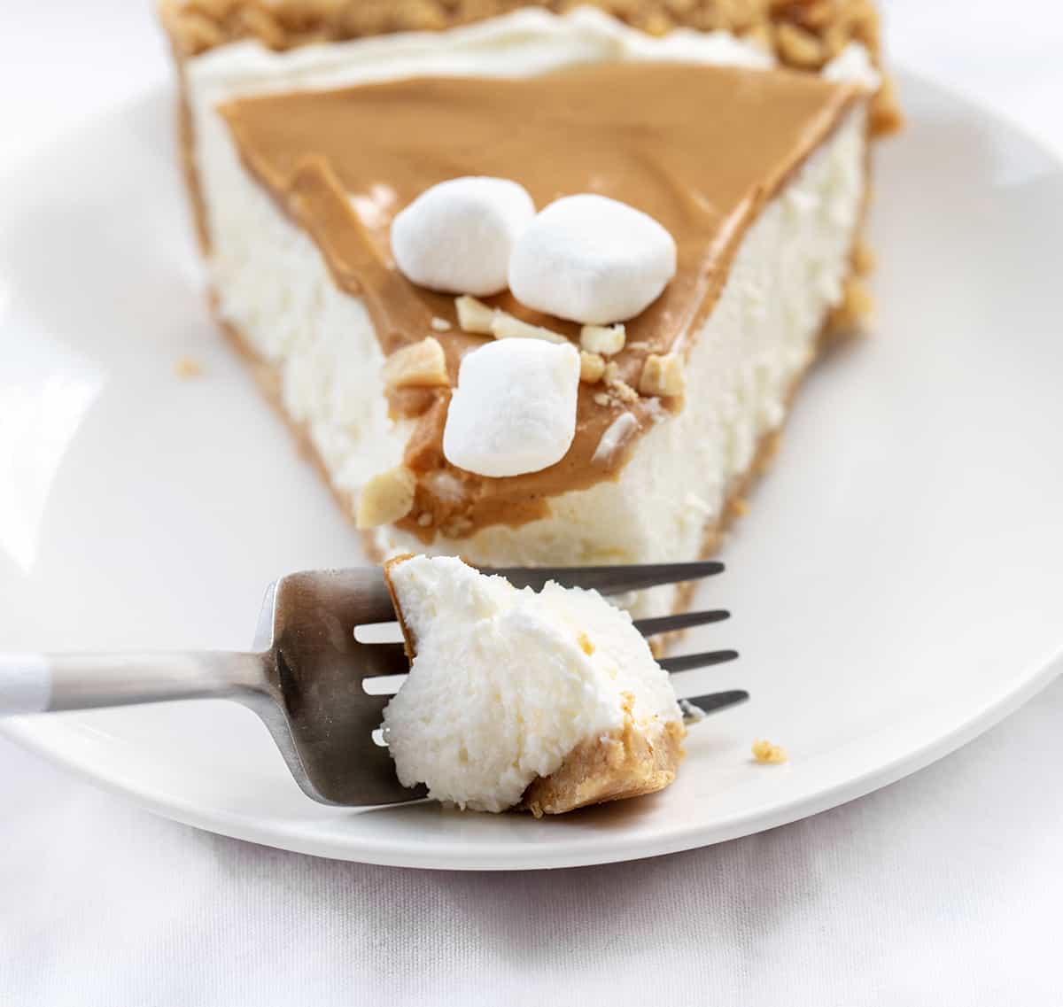Fluffernutter Pie is a no-bake dessert made with a soft and smooth marshmallow filling topped with creamy peanut butter, all in a graham cracker crust. It's another delightful addition to my list of fluffernutter treats that started with my Fluffernutter Cookies! Fork Taking Bite from One Piece of Fluffernutter Pie on White Plate.