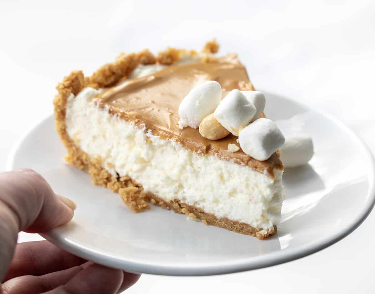 Fluffernutter Pie is a no-bake dessert made with a soft and smooth marshmallow filling topped with creamy peanut butter, all in a graham cracker crust. It's another delightful addition to my list of fluffernutter treats that started with my Fluffernutter Cookies! Hand Holding White Plate with Fluffernutter Pie on It.
