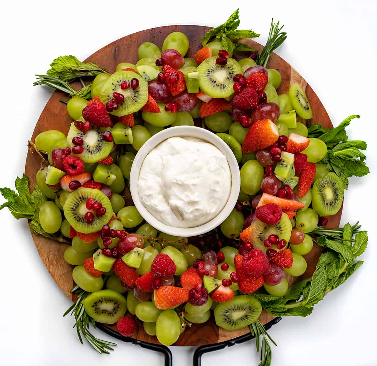 Holiday Wreath Fruit Tray is a beautiful and colorful display of fruit and herbs in a circular design with fruit dip in the center for serving. The fruit tray is presented to represent a wreath, a popular symbol of Christmas. Be sure to add this to your holiday menu! A Holiday Wreath Fruit Tray on a Round Cutting Board.
