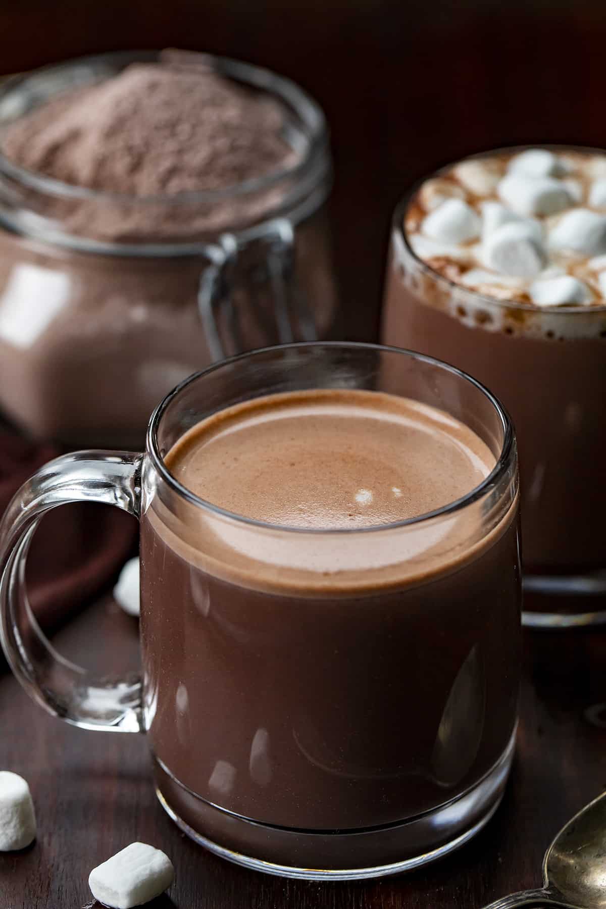 Homemade Hot Cocoa Mix is cheaper than store-bought and tastes so much better! Nothing warms a body up like a mug of rich hot cocoa, especially in the winter. And, it's a beverage that can be enjoyed by all ages. I love that you know exactly what is going into the powdery mix with no added preservatives. This hot chocolate recipe is rich, velvety, decadent, and the perfect beverage for a crowd! Glasses of prepared hot chocolate made from a homemade hot cocoa mix with the jar of the mixture in the background.