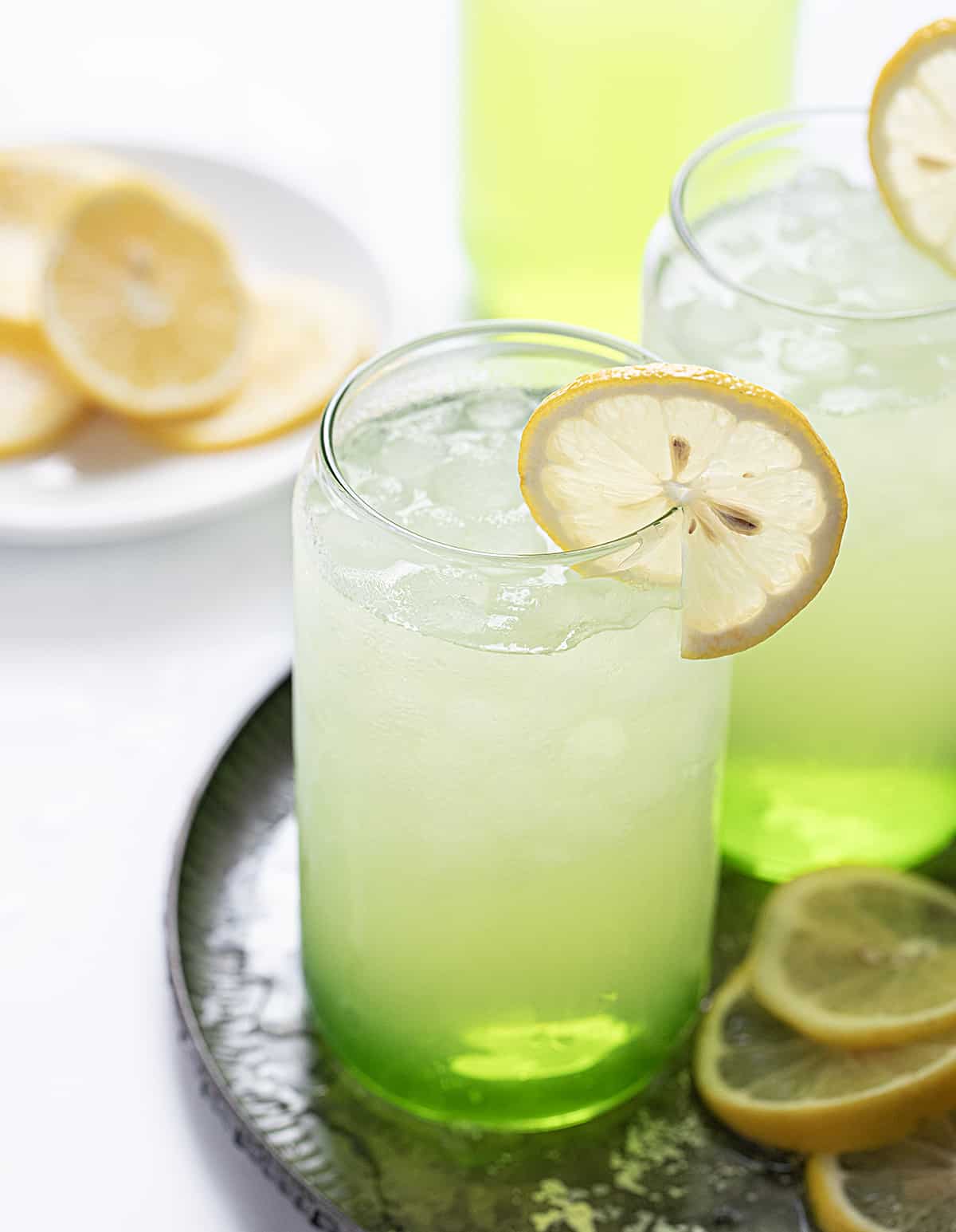 Hulk Smash is an alcoholic beverage made with Midori, sour apple liqueur, and lemonade, all poured over lemonade ice cubes. Both the name and the green color of the drink will remind you of the fictional character 'The Hulk' from Marvel Comics! But don't worry, you won't turn into the Hulk after drinking one, but the sweet drink does pack a punch (pun intended)! Hulk Smash Cocktails on a Tray with Lemon Slices.