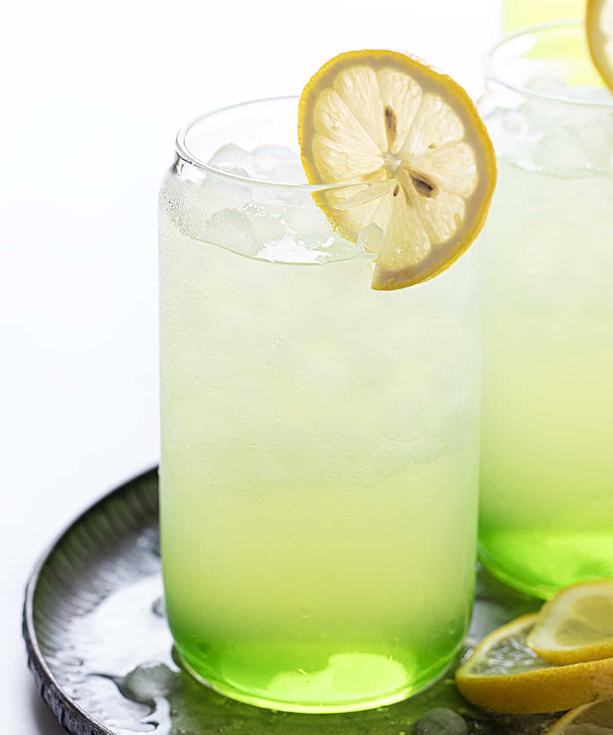 Hulk Smash is an alcoholic beverage made with Midori, sour apple liqueur, and lemonade, all poured over lemonade ice cubes. Both the name and the green color of the drink will remind you of the fictional character 'The Hulk' from Marvel Comics! But don't worry, you won't turn into the Hulk after drinking one, but the sweet drink does pack a punch (pun intended)! Hulk Smash Cocktails on a Tray with Lemon Slices.