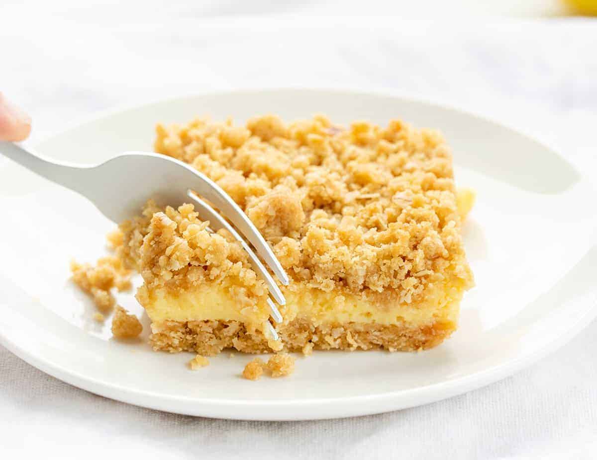Lemon Crumb Bars are dessert bars filled with lemon filling and flavor in between two layers of an oatmeal crust. They are one of the BEST lemon desserts I have ever had! Lemon Bars, Lemon Crumble, Lemon Crumb Bars, Best Lemon Desserts, Dessert, Summer Dessert, Spring Dessert, Easy Dessert, Crowd Pleasing Dessert, Crumble Dessert, recipes, baking, i am baker, iambaker Fork Taking a Bite of Lemon Crumb Bar on a White Plate