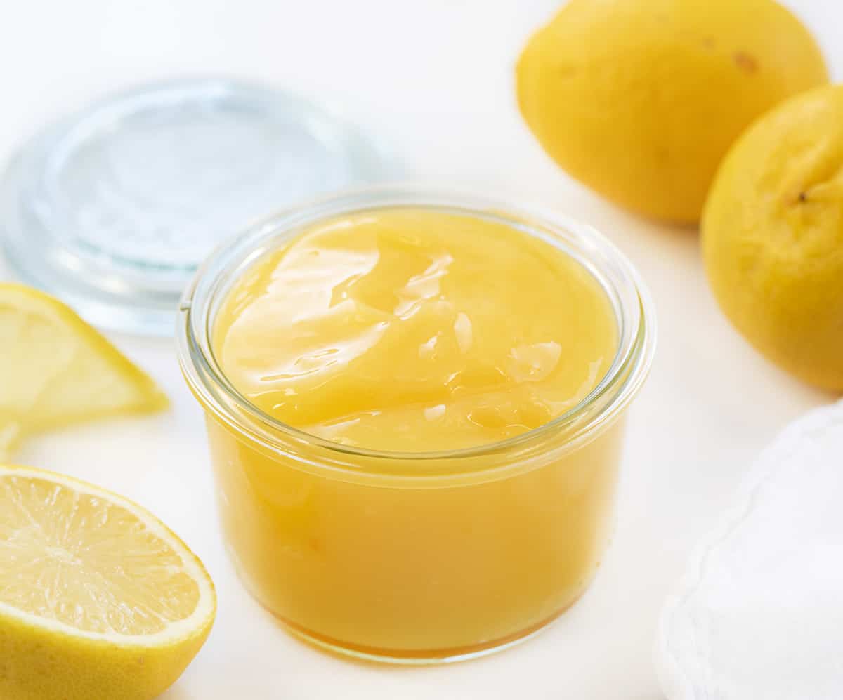 Homemade Lemon Curd is a tart and lemony spread, filling, or topping made from a few simple ingredients. It has a thick and creamy consistency that adds a burst of citrus flavor to many treats and desserts! Jar of Lemon Curd next to lemons.
