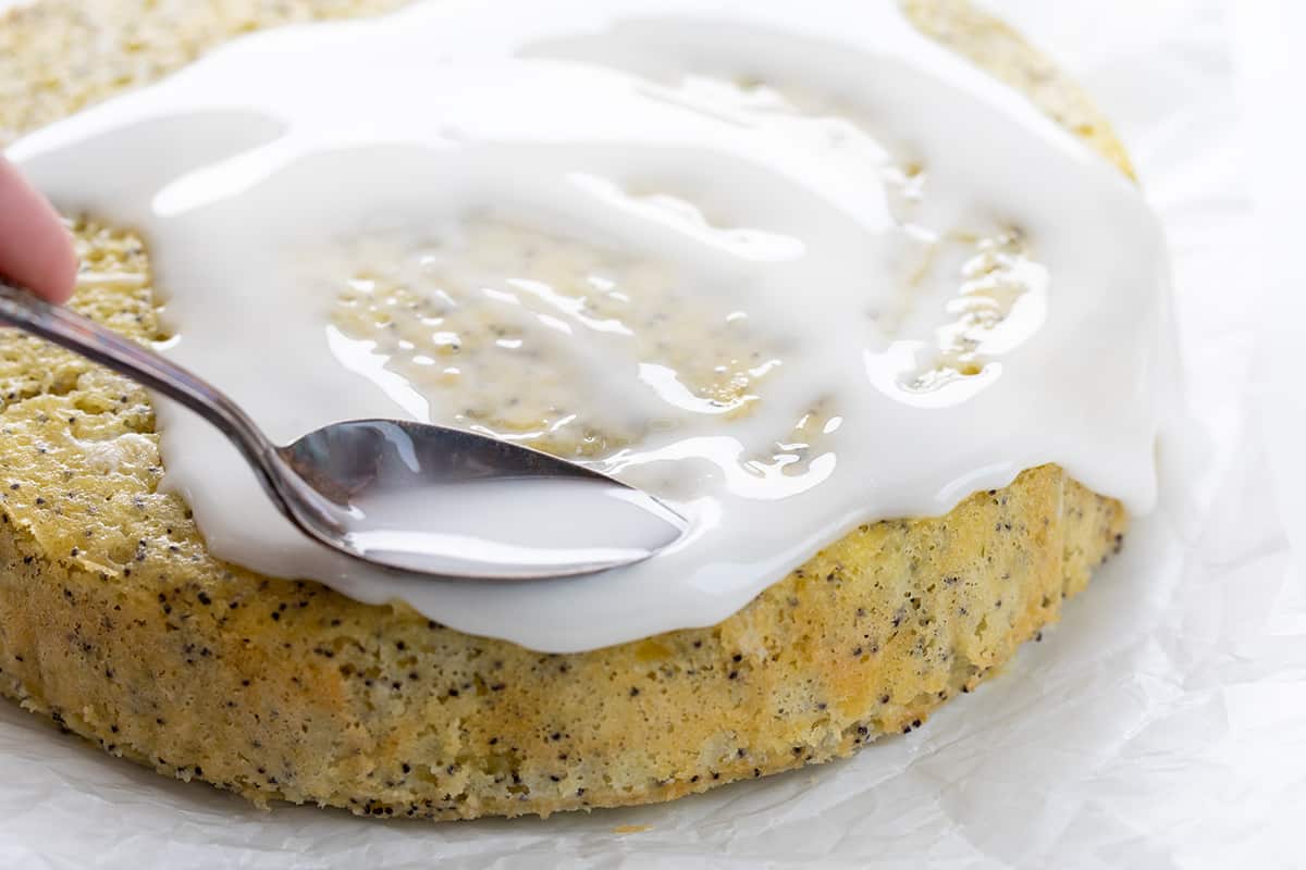Lemon Poppy Seed Cake is a moist and tender lemon-flavored cake with a subtle crunch and nutty undertone from poppy seeds, drizzled with a sweet glaze. A warm milk mixture poured over the cake after it has been baked results in a super soft and delicate crumb. Enjoy this cake with your morning coffee or tea, as a snack, or as a dessert! Spreading Glaze Over Lemon Poppyseed Cake.