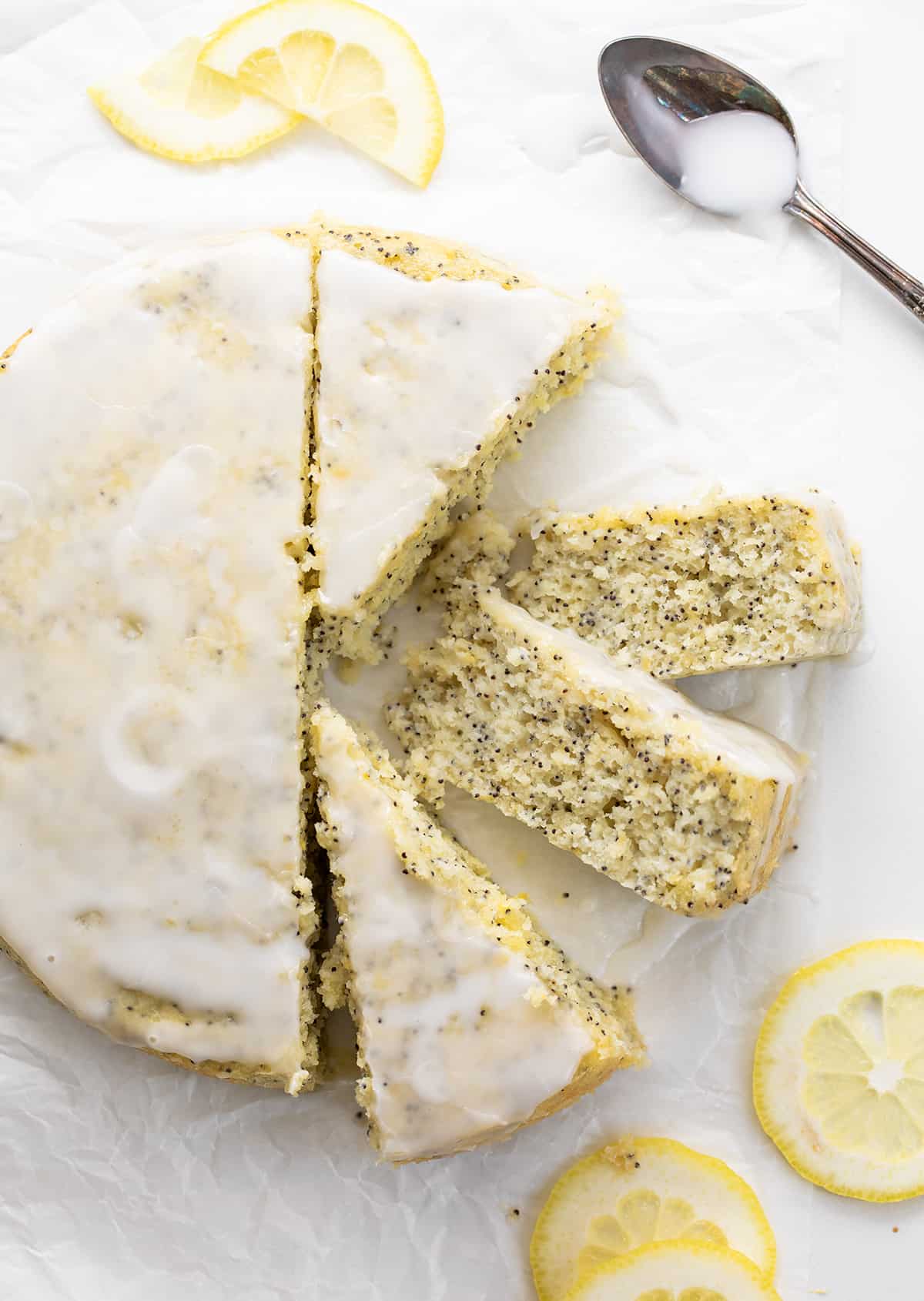 Lemon Poppy Seed Cake is a moist and tender lemon-flavored cake with a subtle crunch and nutty undertone from poppy seeds, drizzled with a sweet glaze. A warm milk mixture poured over the cake after it has been baked results in a super soft and delicate crumb. Enjoy this cake with your morning coffee or tea, as a snack, or as a dessert! Whole Lemon Poppy Seed Cake on a White Piece of parchment Paper with Four Pieces Cut and Two Pieces on Their Sides Showing Tender Crumb.