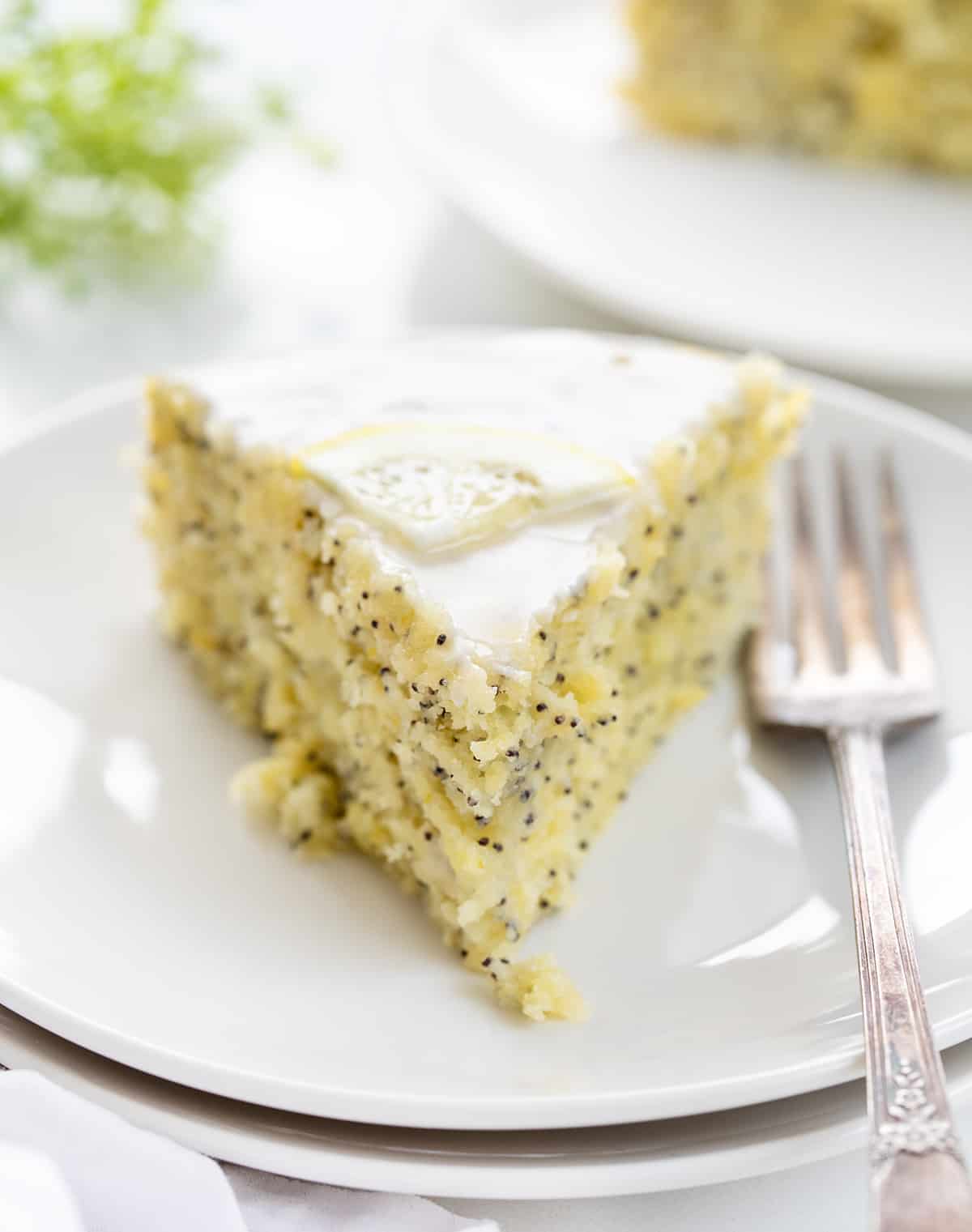 Lemon Poppy Seed Cake is a moist and tender lemon-flavored cake with a subtle crunch and nutty undertone from poppy seeds, drizzled with a sweet glaze. A warm milk mixture poured over the cake after it has been baked results in a super soft and delicate crumb. Enjoy this cake with your morning coffee or tea, as a snack, or as a dessert! Piece of Lemon Poppy Seed Cake on a White Plate with Green Babysbreath in the Background.