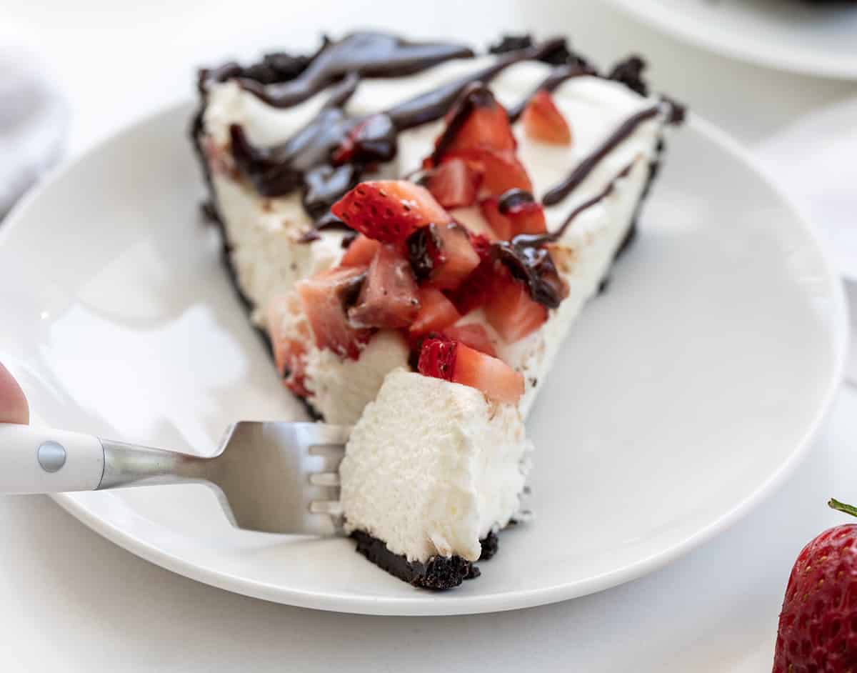 Chocolate Strawberry Marshmallow Pie is a creamy and delicious pie with an Oreo cookie crust filled with a fluffy marshmallow mixture. Then it is casually topped with chopped fresh strawberries and hot fudge! It is a no-bake dessert that would be delightful year-round, but especially in the summertime with fresh strawberries! Fork Taking a Bite of Chocolate Strawberry Marshmallow Pie off a White Plate.