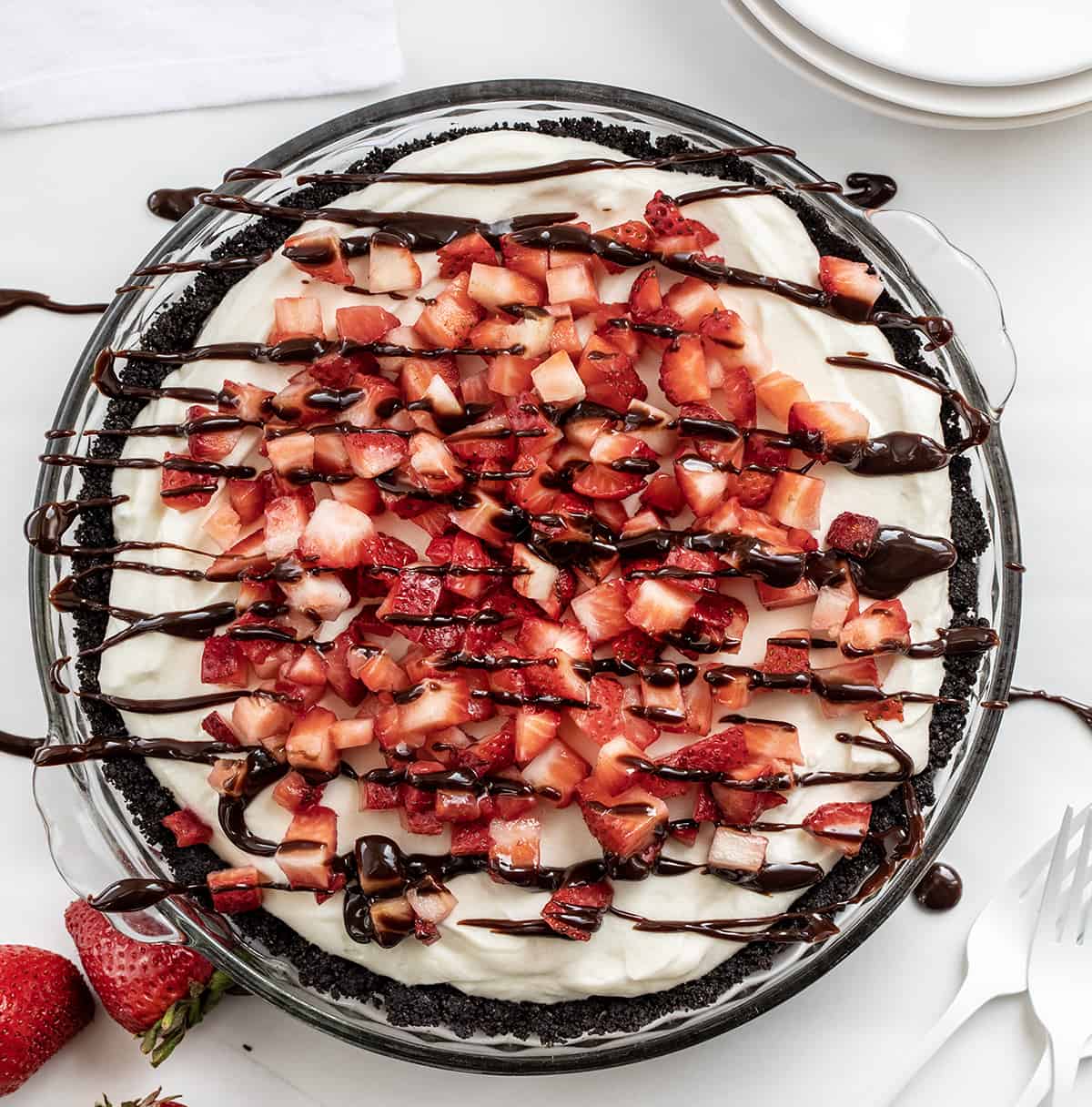 Chocolate Strawberry Marshmallow Pie is a creamy and delicious pie with an Oreo cookie crust filled with a fluffy marshmallow mixture. Then it is casually topped with chopped fresh strawberries and hot fudge! It is a no-bake dessert that would be delightful year-round, but especially in the summertime with fresh strawberries! Overhead of a Chocolate Strawberry Marshmallow Pie on a White Counter with Hot Fudge Drizzle and Strawberries.
