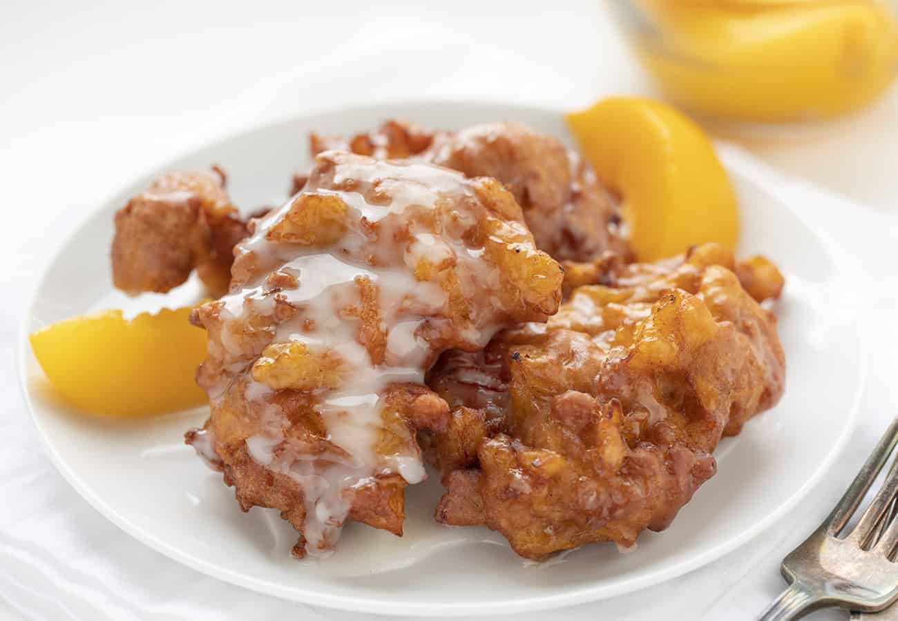 Peach Fritters are made with a cakey batter incorporated with peaches and topped off with a peach-flavored glaze. peach fritters, fritters recipes, homemade glaze, i am baker, iambaker, deep-fried dough, fruit fritters Homemade Peach Fritters on a White Plate with Peaches
