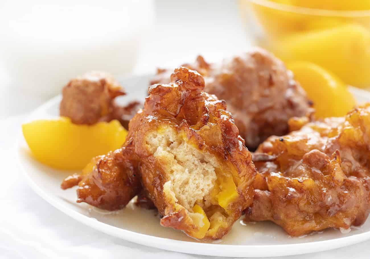 Peach Fritters are made with a cakey batter incorporated with peaches and topped off with a peach-flavored glaze. peach fritters, fritters recipes, homemade glaze, i am baker, iambaker, deep-fried dough, fruit fritters Peach Fritters on a White Plate, one of them bit into