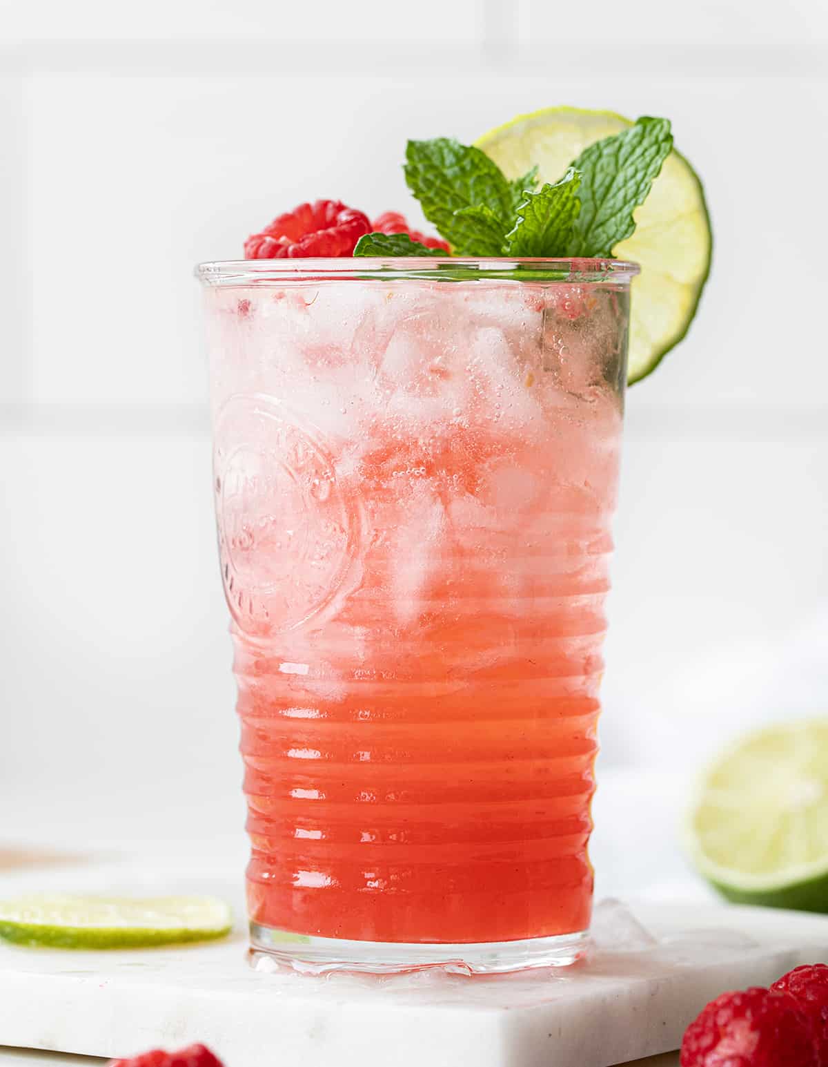 Raspberry Lime Mojito is a refreshing cocktail that combines the flavors of fresh raspberries, zesty lime, light rum, and mint. It is a drink that is perfect for many occasions, especially a hot summer day, brunch with friends, or enjoying an evening at home! One Glass of Raspberry Lime Mojito on a Mini Marble Cutting Board with Limes and Raspberries Around it.