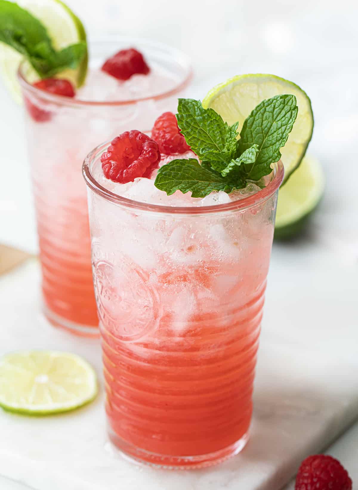 Raspberry Lime Mojito is a refreshing cocktail that combines the flavors of fresh raspberries, zesty lime, light rum, and mint. It is a drink that is perfect for many occasions, especially a hot summer day, brunch with friends, or enjoying an evening at home! Two Glasses of Raspberry Lime Mojito on a Mini Marble Cutting Board with Limes and Raspberries Around it.
