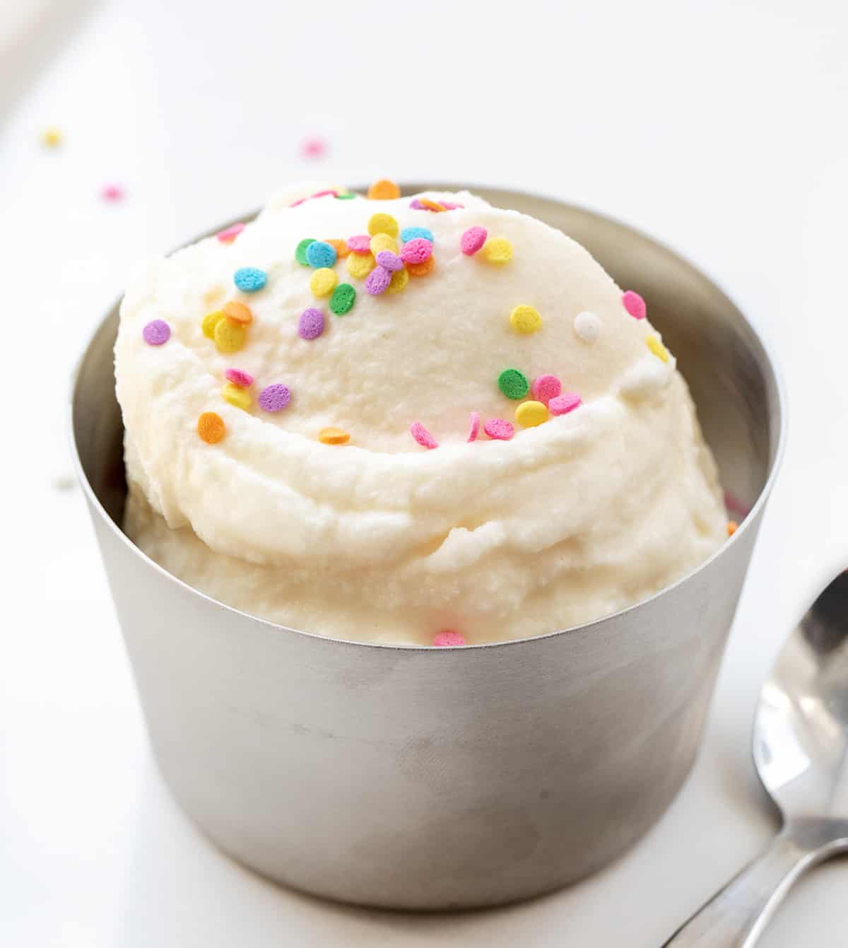 Quick and Easy Soft Serve Ice Cream (also known as small batch ice cream in a bag!)is smooth and creamy vanilla ice cream made with no special equipment and ready in about 5 minutes! All you need is a little arm strength, and then the ice cream will be ready to enjoy! This is a wonderful recipe for kids to be involved! Metal Bowl with Scoops of Soft Serve Ice Cream in It with Sprinkles and a Spoon on a White Counter.