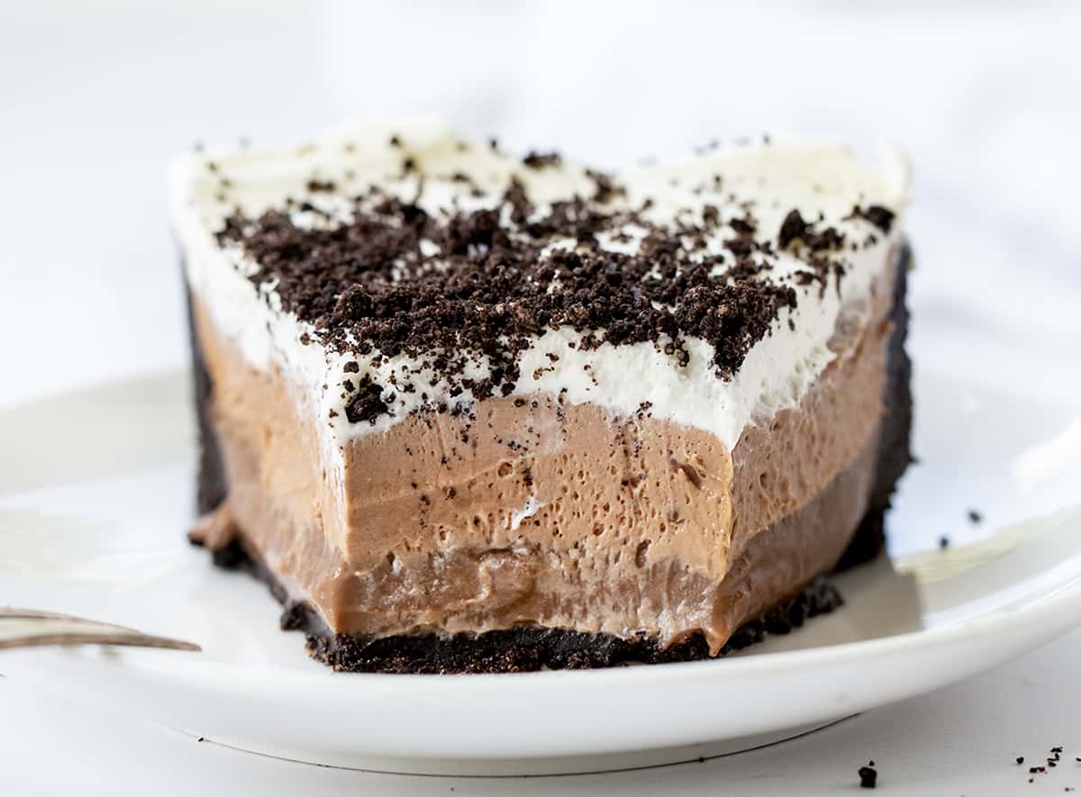 Triple Chocolate Mousse Pie is a decadent no-bake layered dessert with an Oreo crust topped with chocolate pudding, chocolate mousse, and whipped topping. It's a treat that chocolate lovers will savor and enjoy! Piece of Triple Chocolate Mousse Pie on a White Plate with Some Removed Showing Texture.