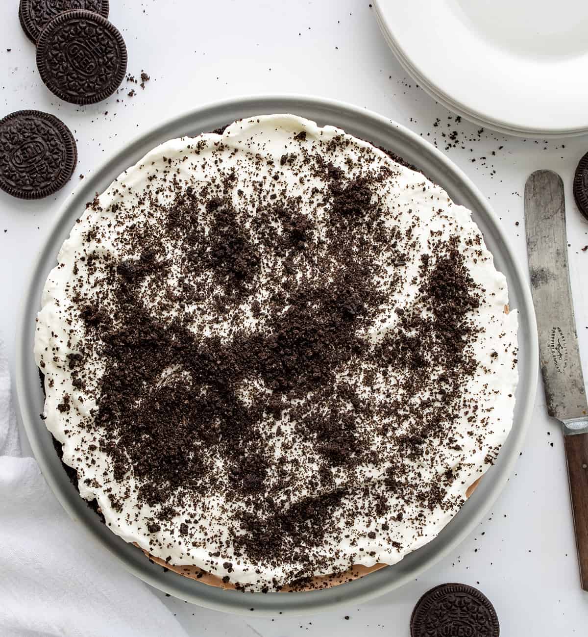 Triple Chocolate Mousse Pie is a decadent no-bake layered dessert with an Oreo crust topped with chocolate pudding, chocolate mousse, and whipped topping. It's a treat that chocolate lovers will savor and enjoy! Overhead of a Triple Chocolate Mousse Pie on a White Counter with Oreos and Plates.