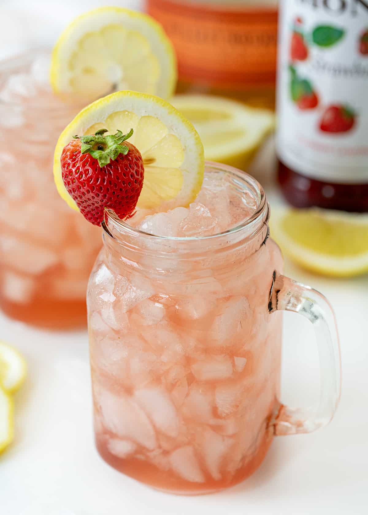 Whiskey Strawberry Lemonade combines the tanginess of lemon juice with the sweetness of strawberry syrup, balanced with a splash of whiskey. It's a refreshing cocktail that is served over crushed ice, garnished with lemon slices and strawberries for the finishing touch. It's as pretty to look at as it is delightful to sip! Glasses of Whiskey Strawberry Lemonade on a White Counter with Strawberry Syrup in the Background.