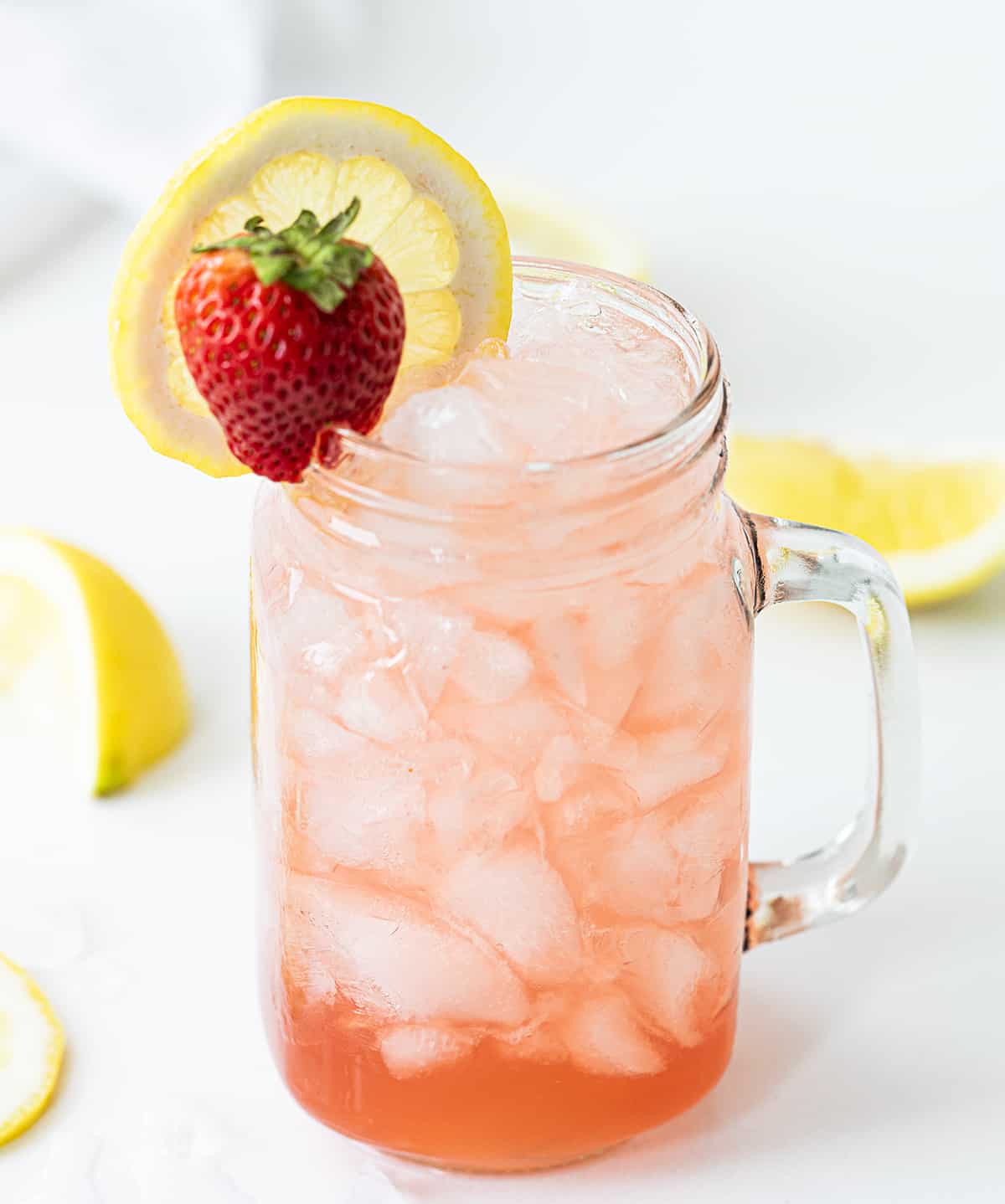 Whiskey Strawberry Lemonade combines the tanginess of lemon juice with the sweetness of strawberry syrup, balanced with a splash of whiskey. It's a refreshing cocktail that is served over crushed ice, garnished with lemon slices and strawberries for the finishing touch. It's as pretty to look at as it is delightful to sip! Glass Mug with Whiskey Strawberry Lemonade and a Strawberry and Lemon Garnish.